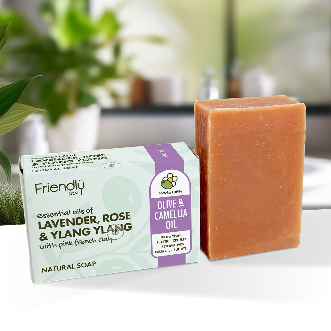 A bar of olive and laveder rose and ylang ylang soap stands next to its packaging box on a white surface. The soap bar is a warm peachy-orange color, and the box is light green and white with purple accents.
