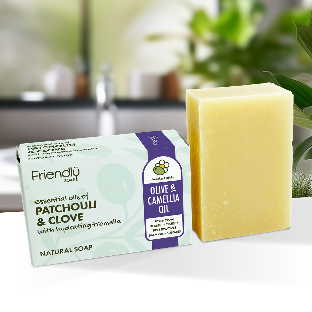 A bar of pale yellow soap stands next to its retail box on a white surface. The box is light green and purple with text reading 'Friendly Soap' and 'Olive & Camellia Oil' and 'Essential Oils of Patchouli & Clove with hydrating tremella.