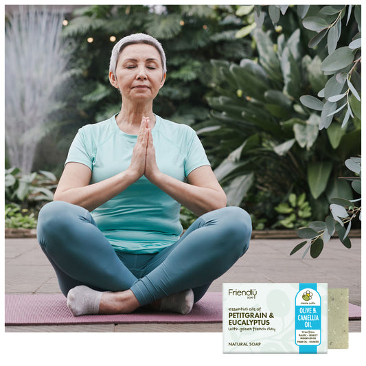 A woman in a light blue t-shirt and teal leggings sits cross-legged on a yoga mat in a garden setting with her hands pressed together in a meditative pose. In the bottom right corner, there is a product packaging overlay for Friendly Natural Soap Petitgrain & Eucalyptus soap.