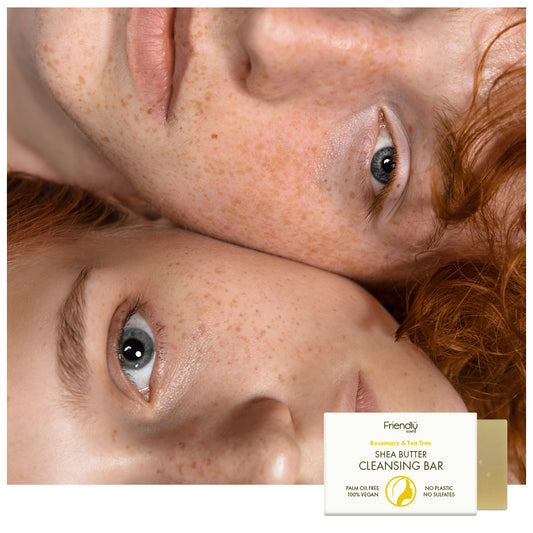 Two close-up faces with freckles and red hair resting together, with a small product box and a cleansing bar at the bottom right. The box reads 'Friendly Rosemary & Tea Tree Shea Butter Cleansing Bar.