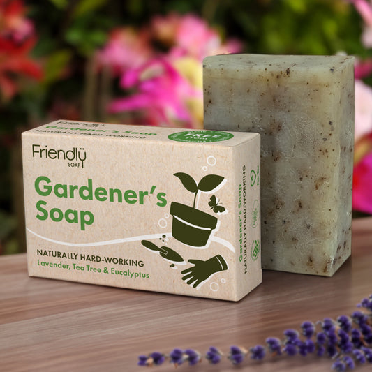 A bar of gardening soap next to its packaging, which is labeled 'Friendly Gardener's Soap' with illustrations of plants and gardening symbols. The bar appears to be made of natural ingredients with exfoliants.