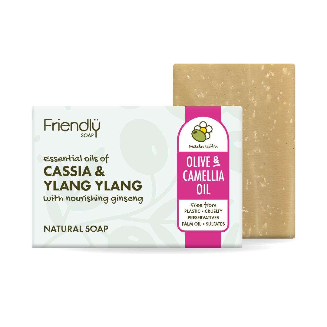 A bar of Friendly soap with its retail packaging, featuring a light beige speckled soap bar behind a white and pink wrapper labeled 'Olive & Camellia Oil'.