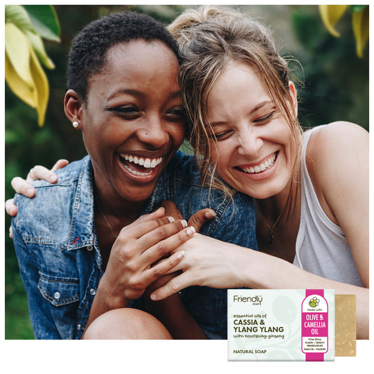 Two smiling women hugging outdoors, with a bar of Friendly brand 'Cassia & Yang Yang' natural soap shown in the bottom right corner.