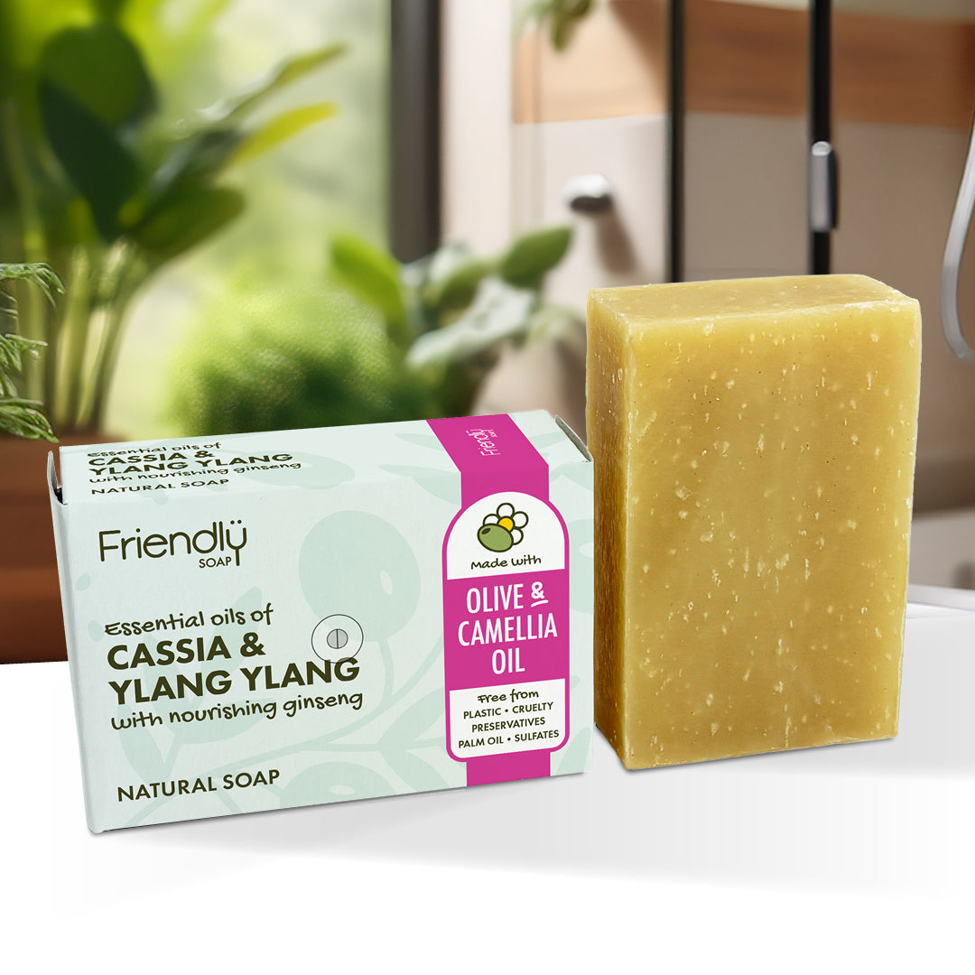 A bar of yellow-green natural soap stands next to its retail box on a white surface. The box reads 'Friendly Soap' and 'Olive & Camellia Oil' with 'Essential Oils of Cassia & Yang Yang'.