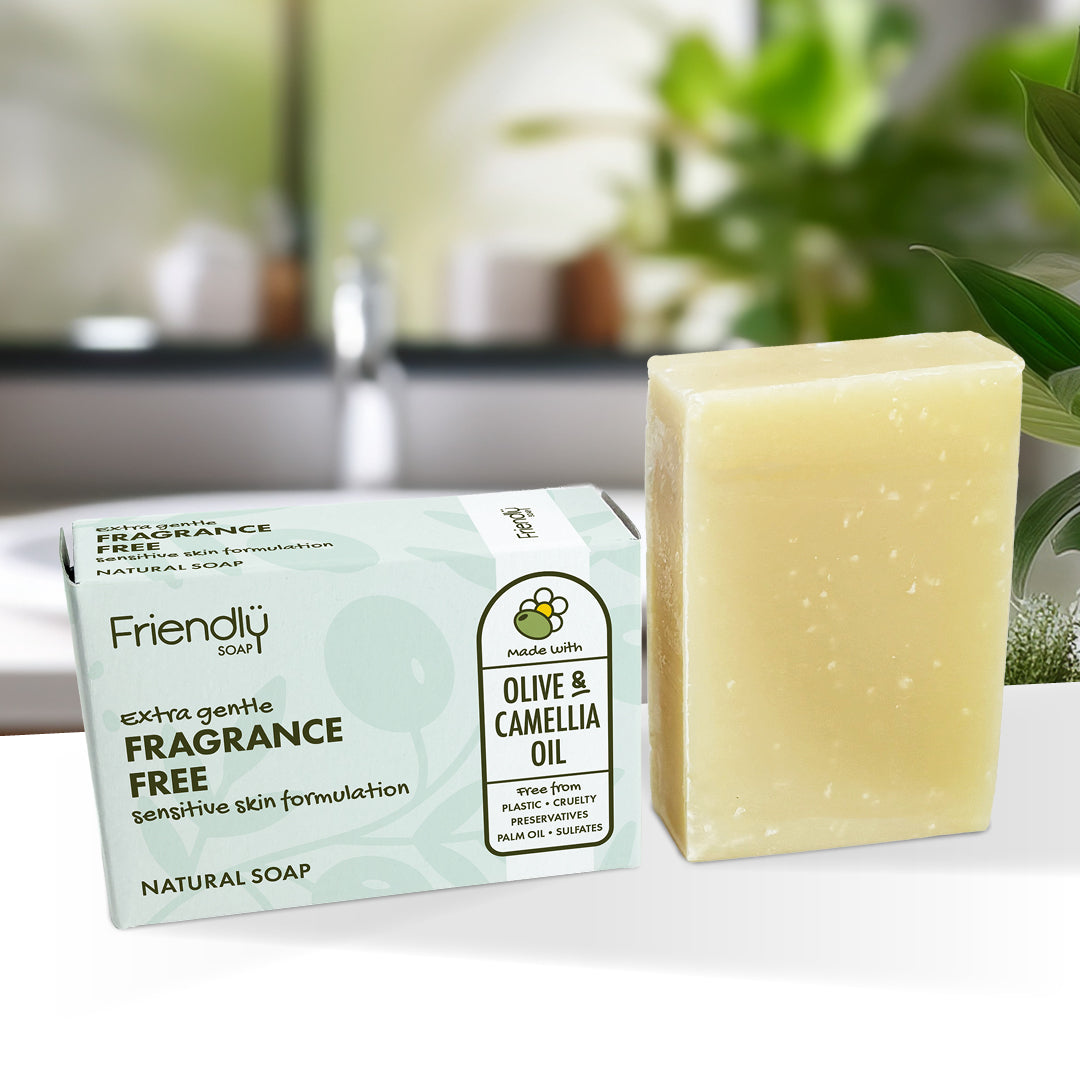 A rectangular bar of pale yellow soap stands beside its mint-green and white box on a white surface, with a softly blurred indoor background and green plants behind it. The box reads Friendly Soap FRAGRANCE FREE Olive & Camellia Oil.