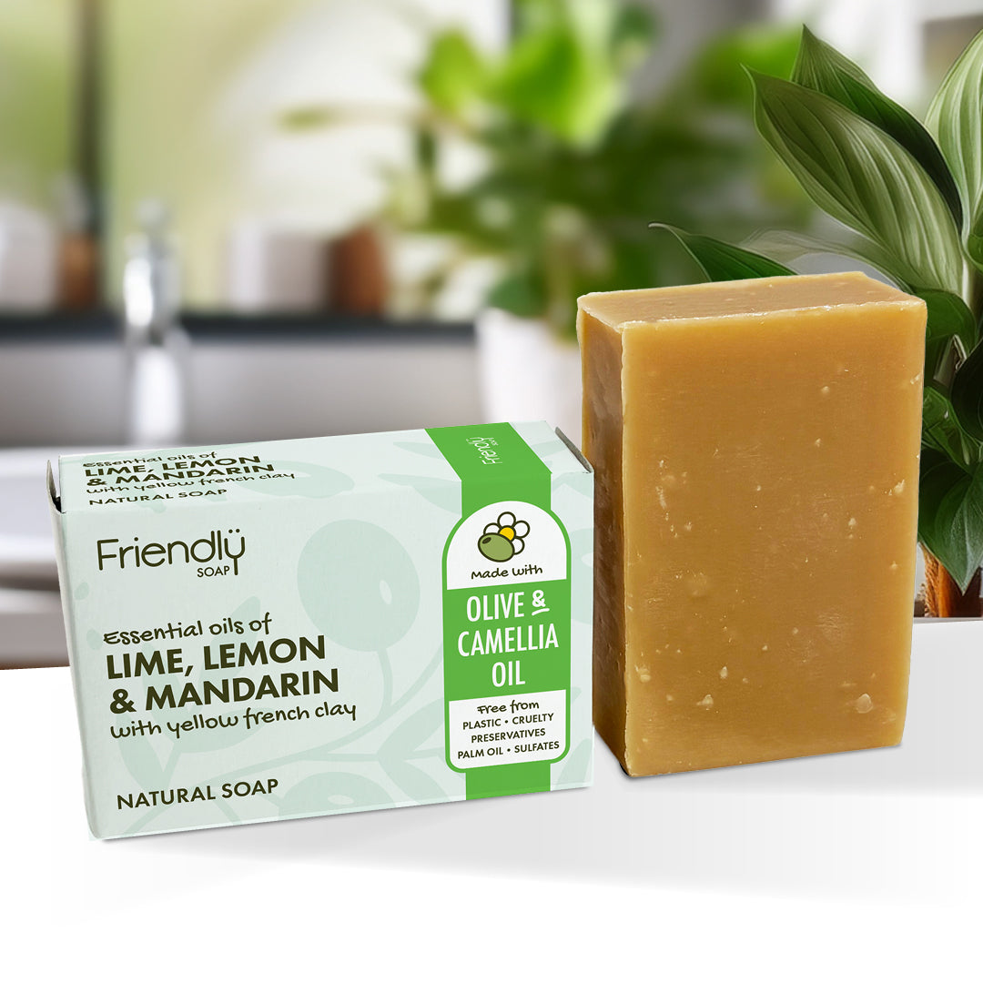 A bar of lime, lemon & mandarin soap made with olive and camellia oil stands next to its packaging box on a white surface, with a blurred kitchen sink and green plants in the background.