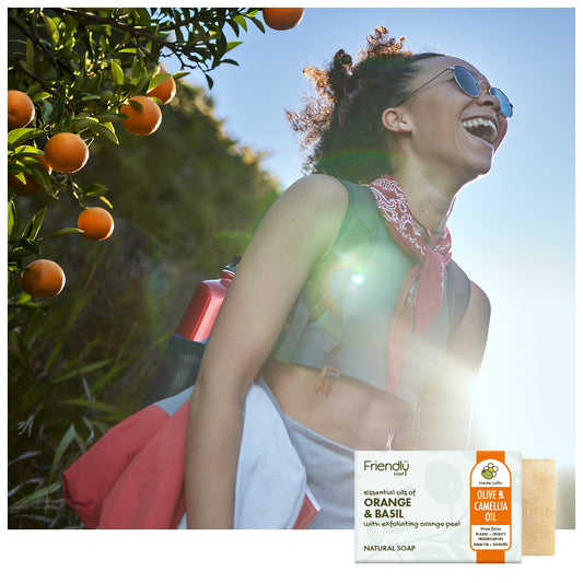 A woman outdoors in a sunny citrus grove wears sunglasses and a pink bandana, with a bar of soap and its packaging shown in the lower right corner. The soap label reads Friendly and Orange & Basil.