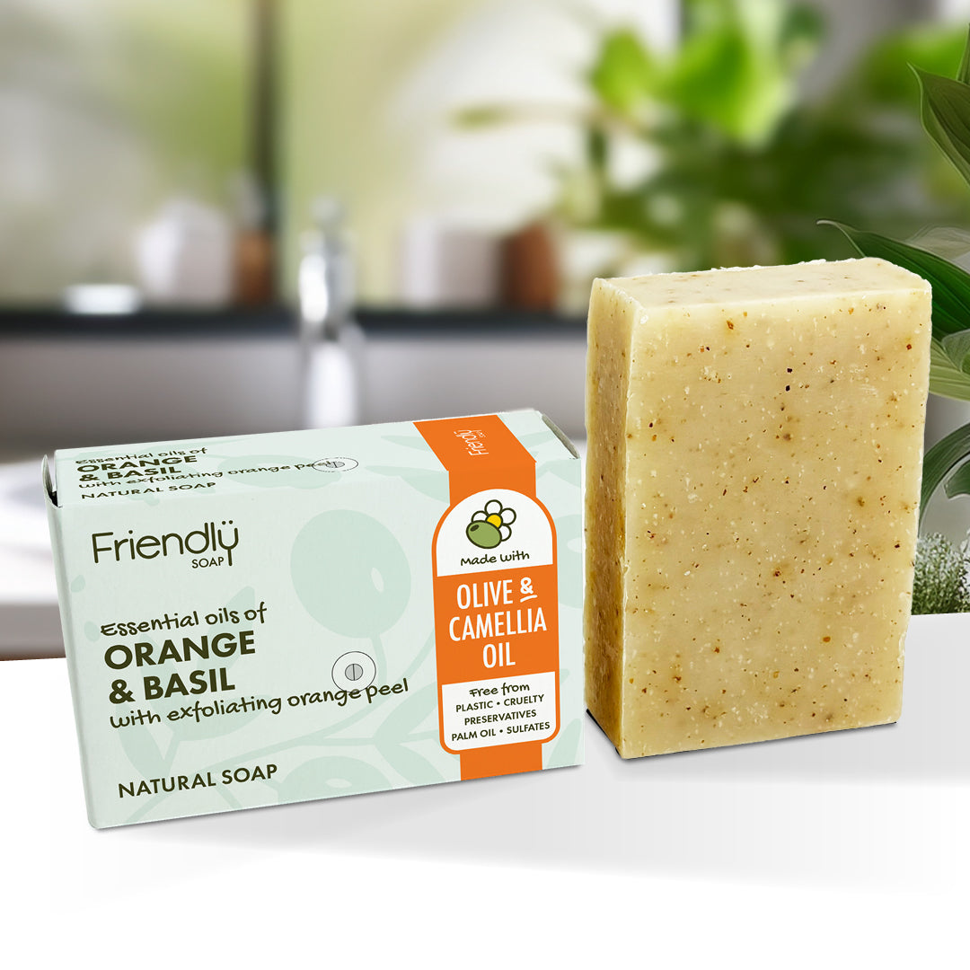 A bar of handmade soap stands next to its retail box on a white surface. The box is light mint green with orange accents and reads 'Friendly Soap' and 'Orange & Basil'.