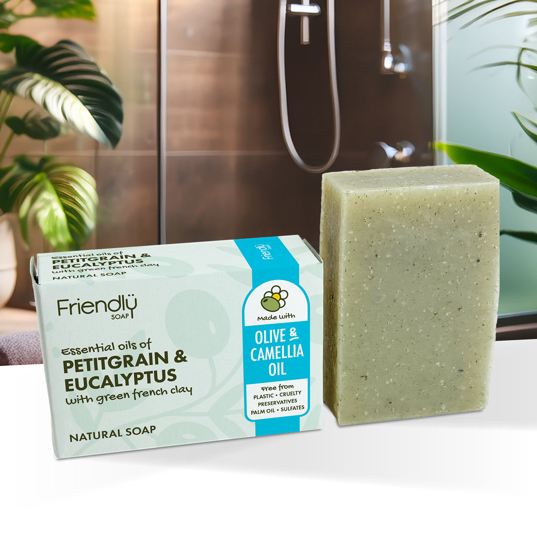 A bar of olive and camellia oil soap stands next to its retail box on a white surface. The box is labeled 'Friendly Soap' and 'Essential oils of Petitgrain & Eucalyptus with green french clay'.