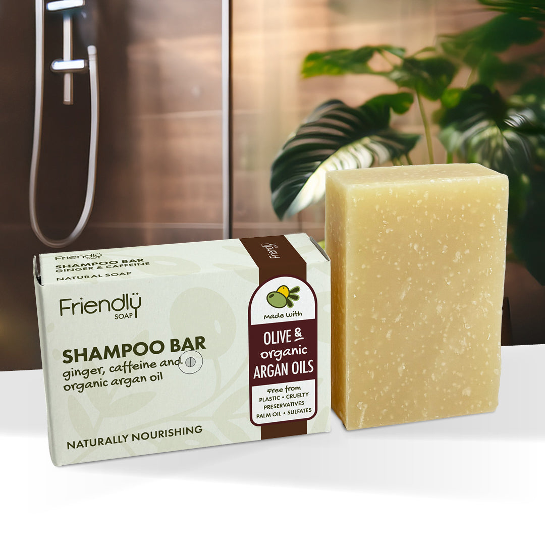 A bar of olive and organic argan oils shampoo bar by Friendly Soap, shown next to its packaging box. The soap bar is a pale yellowish-beige color, and the box is white with brown and green accents, set against a blurred background of a bathroom and green plants.