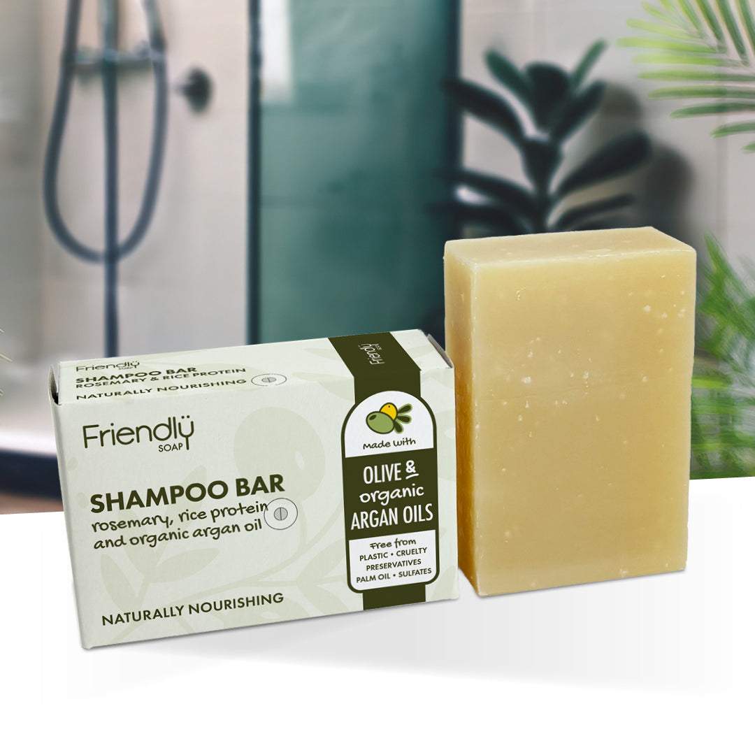 A bar of Friendly Soap shampoo bar next to its packaging box, set on a white surface with a blurred background of a bathroom and green plants.