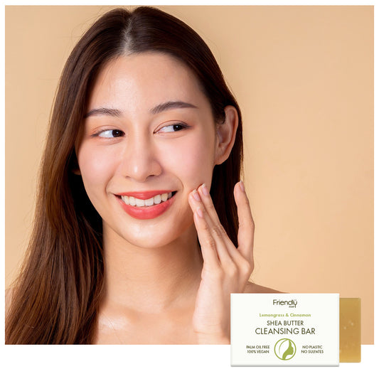 A smiling woman with clear skin touches her cheek beside a box of Friendly Skin Shea Butter Cleansing Bar on a warm beige background.