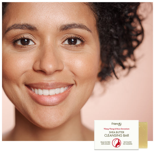 Close-up of a smiling woman with clean, skin next to a small bar of Friendly Soaps cleansing bar packaging on a soft peach background.