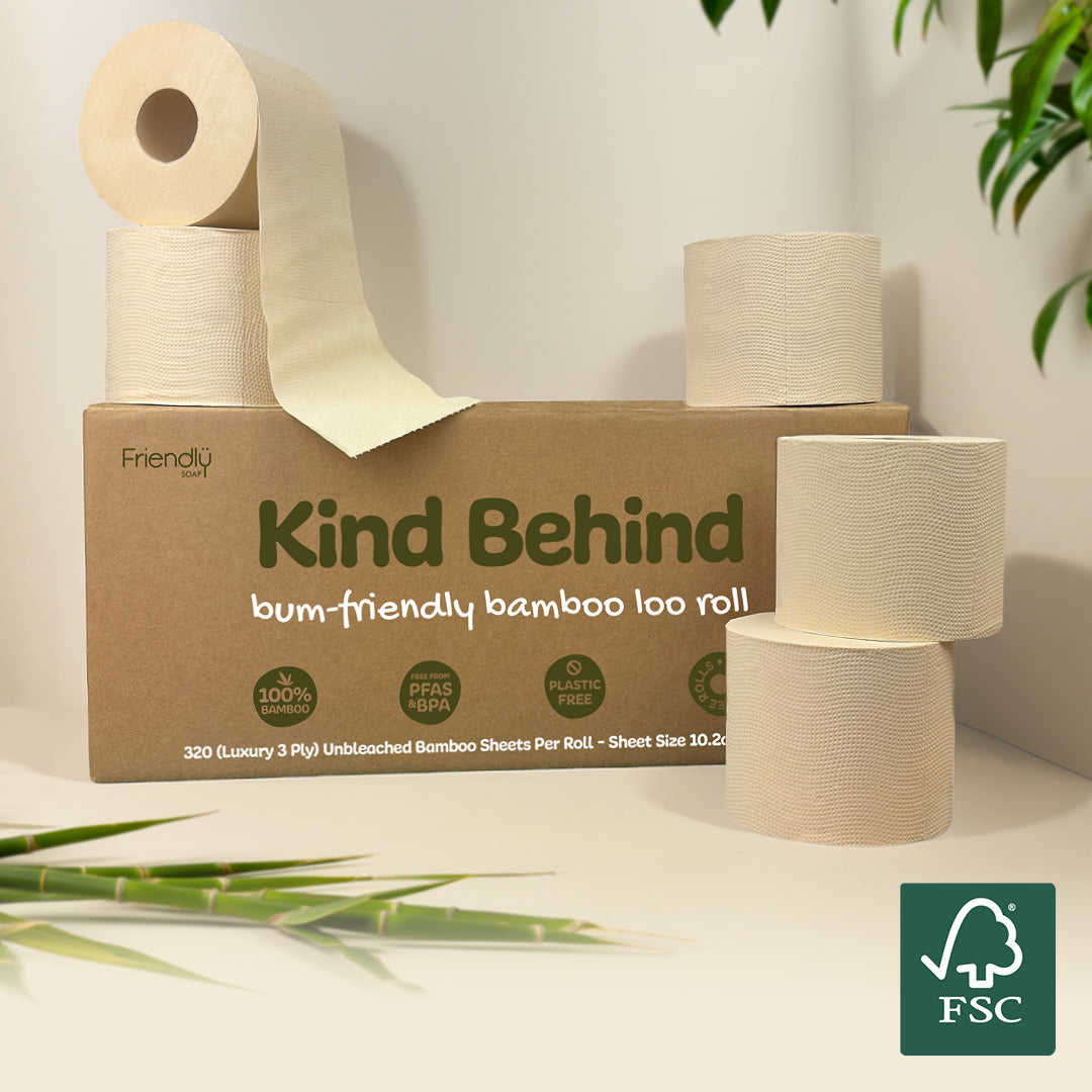 A box of Friendly Branded 'Kind Behind' unbleached bamboo toilet paper rolls, with several rolls displayed around the box on a light beige surface against a white wall, with decorative green bamboo leaves in the corners.