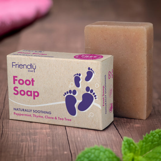 A bar of Foot Soap next to its packaging, which is labeled 'Friendly Soap' with images of feet and spa-like imagery. The bar is beige, and there is mint leaves in the foreground.