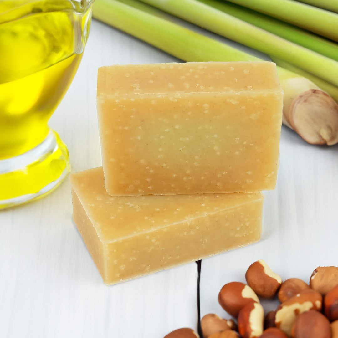 Two rectangular bars of lemongrass handmade soap stacked on a white surface, with lemongrass and yellow oil in the background.