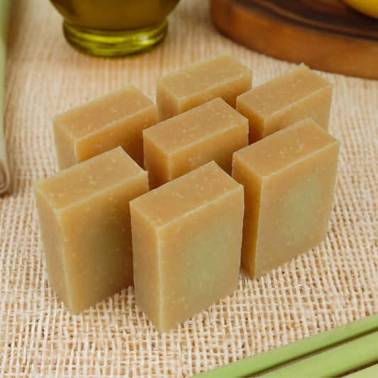 Seven rectangular bars of lemongrass natural soap arranged on a woven beige mat, with a softly blurred olive-green bottle and wooden surface in the background.