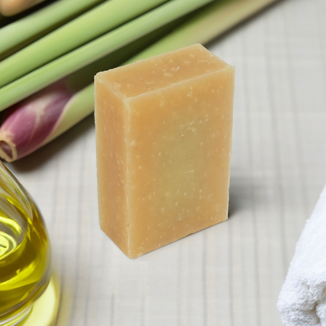 A rectangular bar of pale colored handmade lemongrass natural soap standing upright on a light textured surface, with lemongrass, a bottle of oil, and a white towel in the background.