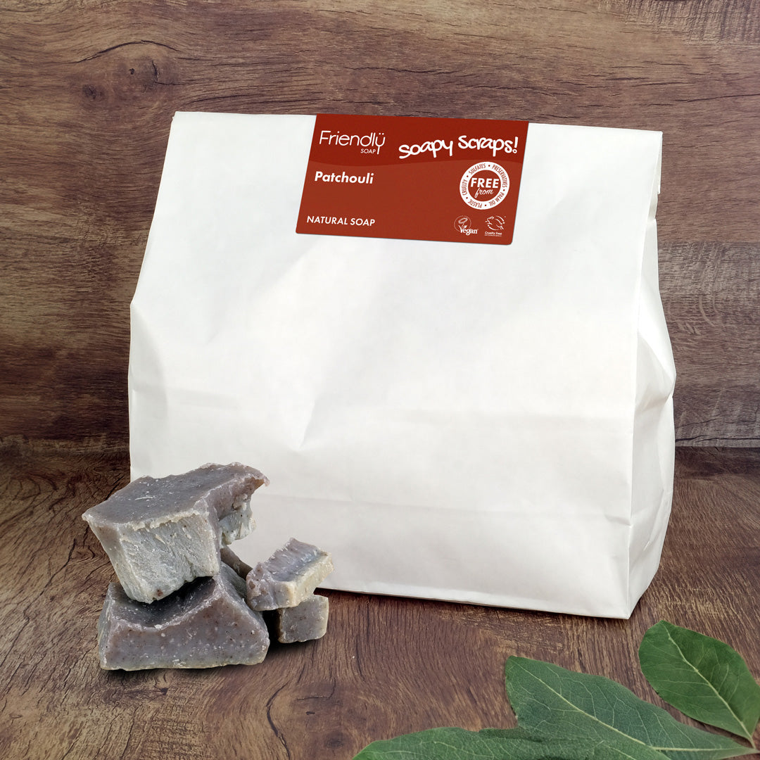 A white paper bag of Friendly Soaps 'Soapy Scraps' Patchouli Natural Soap stands on a wooden surface with several grayish-purple soap bars and green leaves in the foreground.